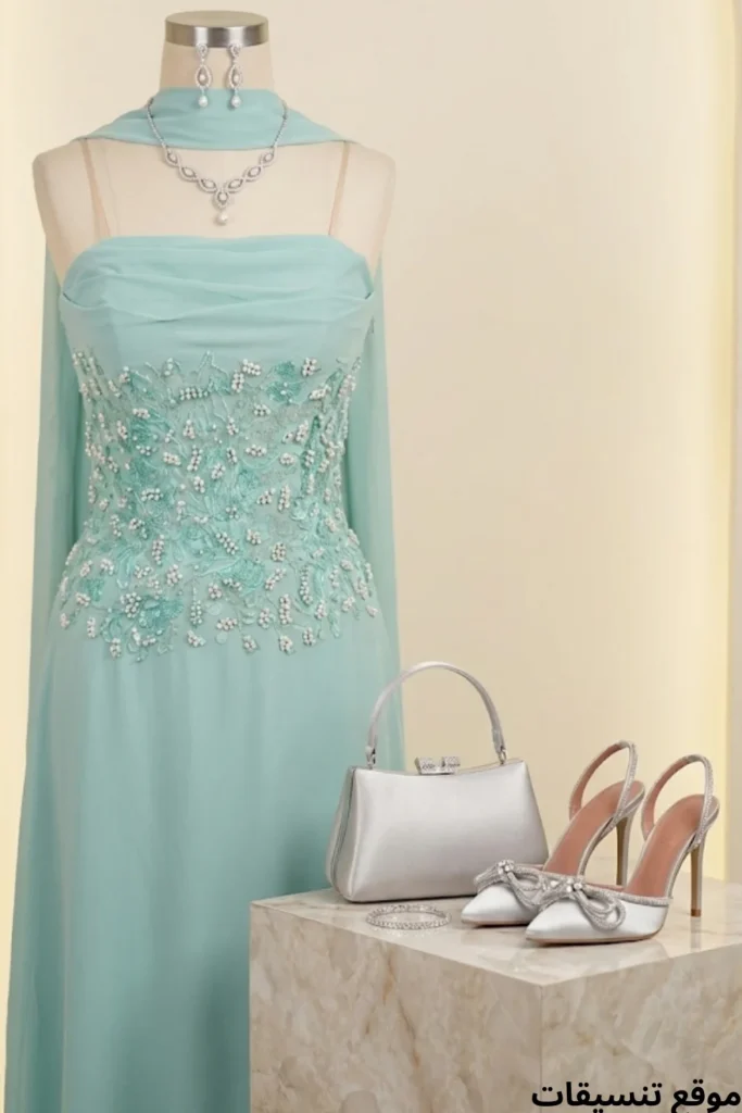 صورة تنسيق فستان سهرة ناعم باللون التركوازي الفاتح مزين بتطريزات لؤلؤ، مع شال متصل، طقم مجوهرات فضي، حقيبة يد فضية، وحذاء فضي بكعب مزين بفيونكة كريستالية.