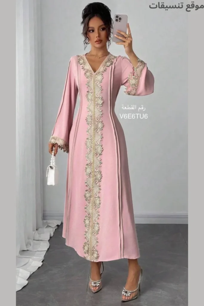 مودل ترتدي جلابية بستايل قفطان مغربي ناعم باللون الوردي، تطريز "سفيفة" طولي من الأعلى للأسفل، أكمام واسعة، وحقيبة بيضاء صغيرة.